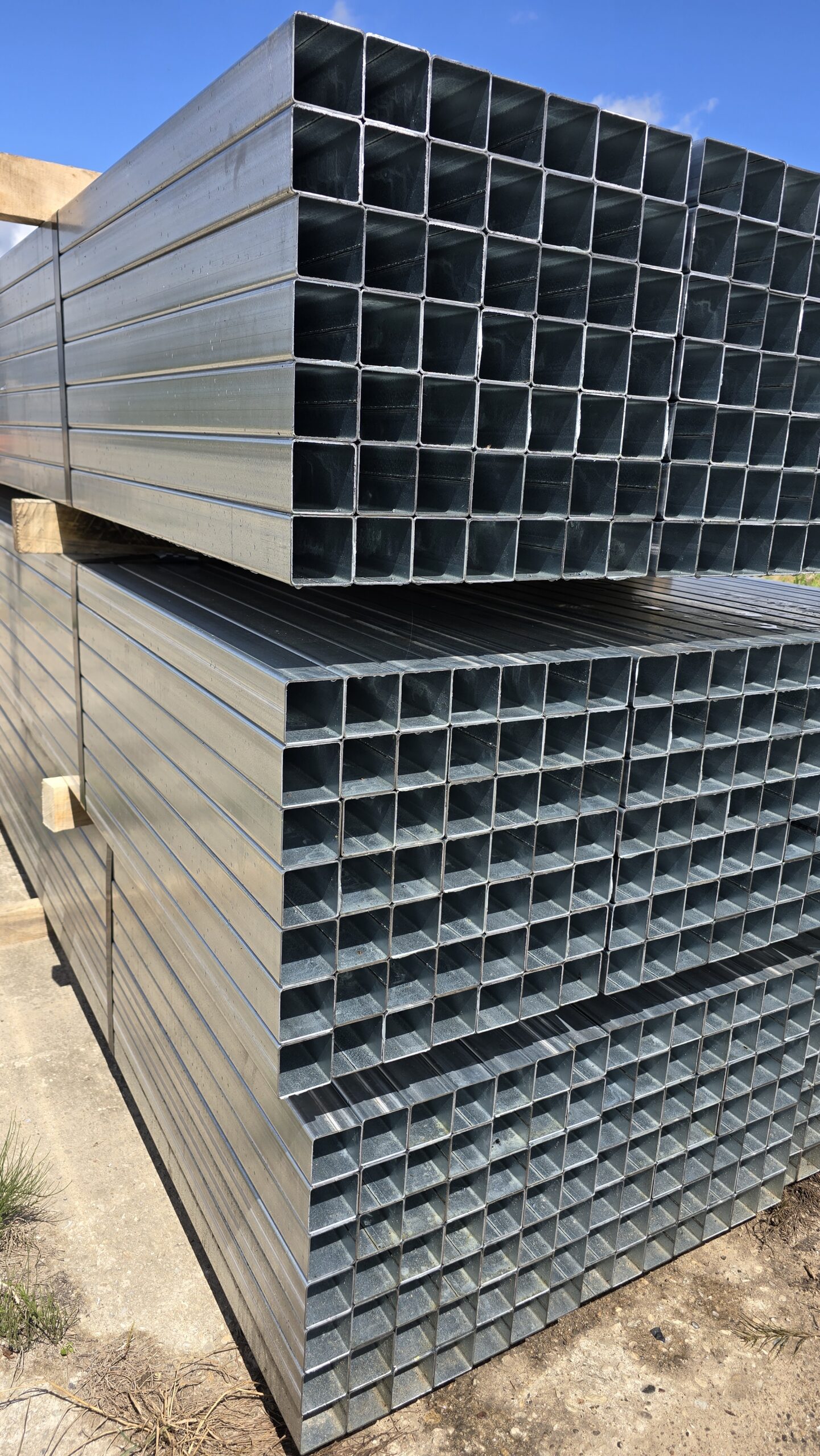 Bundles of galvanized square steel tubing stacked on timber dunnage outdoors.